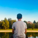 A young man sitting alone facing a still pond, seen from behind, reflecting in quiet solitude