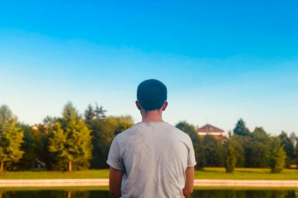 A young man sitting alone facing a still pond, seen from behind, reflecting in quiet solitude