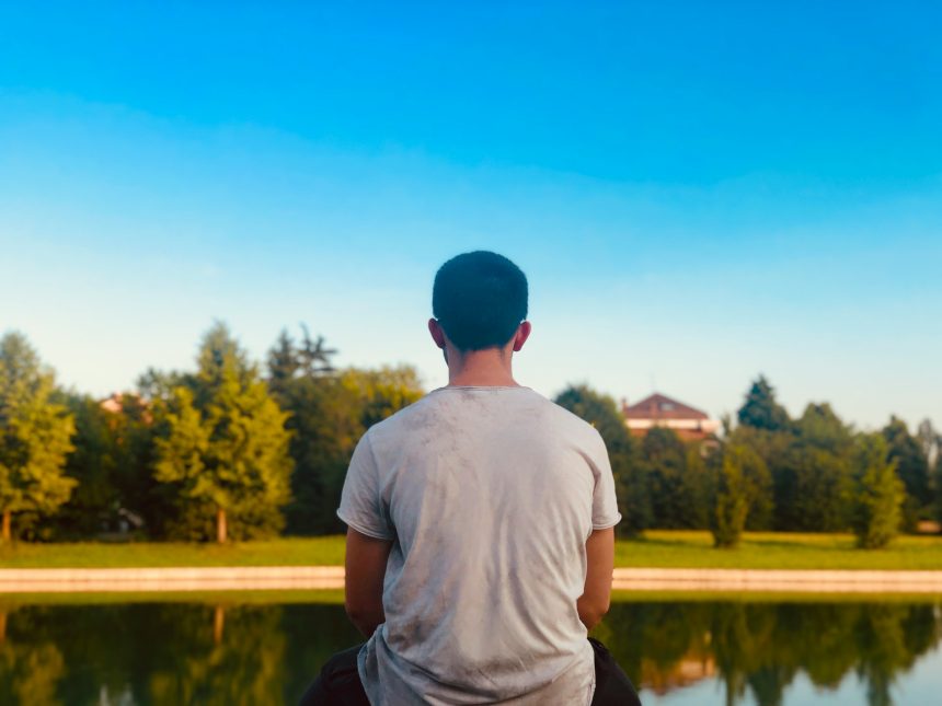 A young man sitting alone facing a still pond, seen from behind, reflecting in quiet solitude