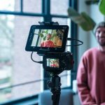 A content creator filming himself at a desk with a camera and monitor in a home studio setup