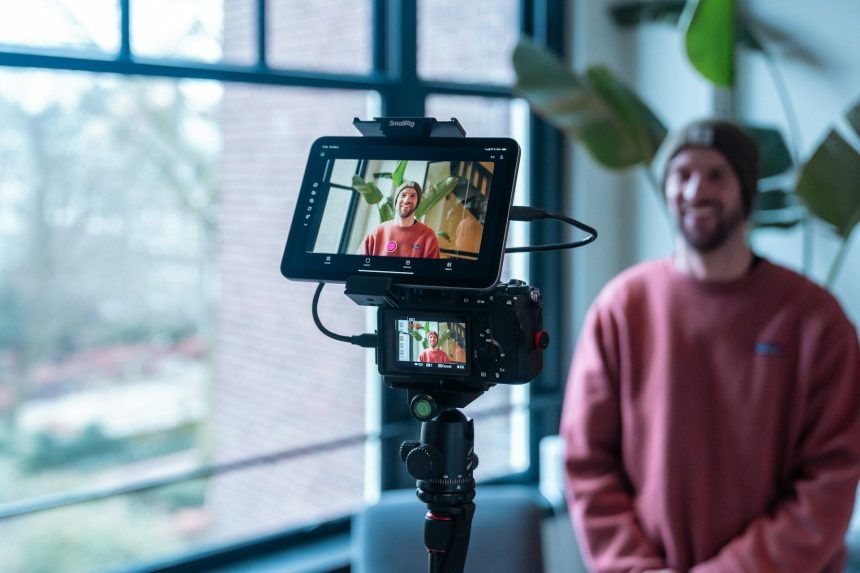 A content creator filming himself at a desk with a camera and monitor in a home studio setup