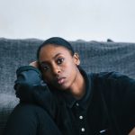 A young Black woman sitting alone on the floor, leaning against a grey sofa, with a quiet and contemplative expression