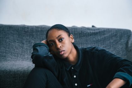 A young Black woman sitting alone on the floor, leaning against a grey sofa, with a quiet and contemplative expression