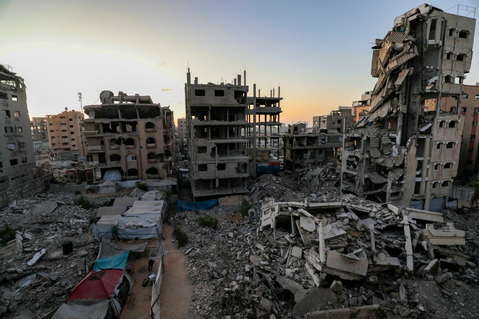 Bombed-out residential buildings stand gutted against a warm sunset sky, with rubble and makeshift shelters filling the foreground, a scene of civilisational collapse in the midst of lingering beauty.