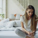 Woman sitting quietly on bed, partner sleeping behind her, early morning light