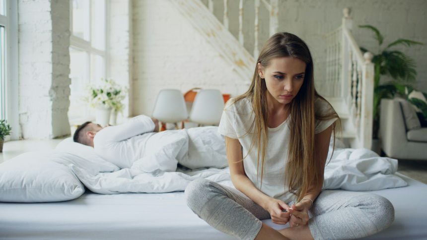 Woman sitting quietly on bed, partner sleeping behind her, early morning light