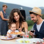 A multigenerational family gathered around an outdoor table at a garden barbecue, sharing a meal together in warm natural light.