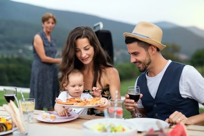 A multigenerational family gathered around an outdoor table at a garden barbecue, sharing a meal together in warm natural light.