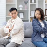A couple sitting back to back on a sofa with arms crossed, showing emotional distance and lack of communication.