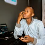 Young Nigerian man using laptop and phone in a modest indoor setting
