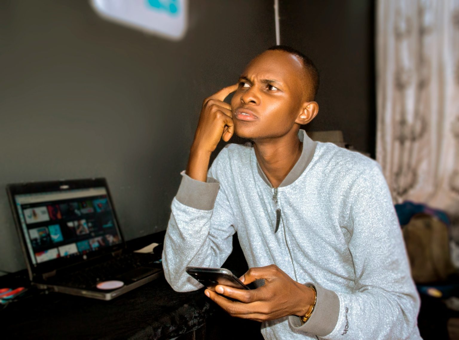 Young Nigerian man using laptop and phone in a modest indoor setting
