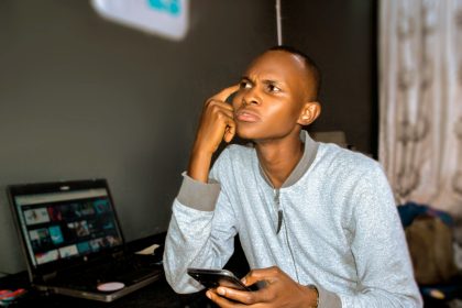 Young Nigerian man using laptop and phone in a modest indoor setting